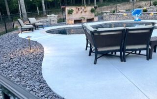 Finished backyard pool area with concrete patio, dining set, lounge chairs, and outdoor fireplace surrounded by trees