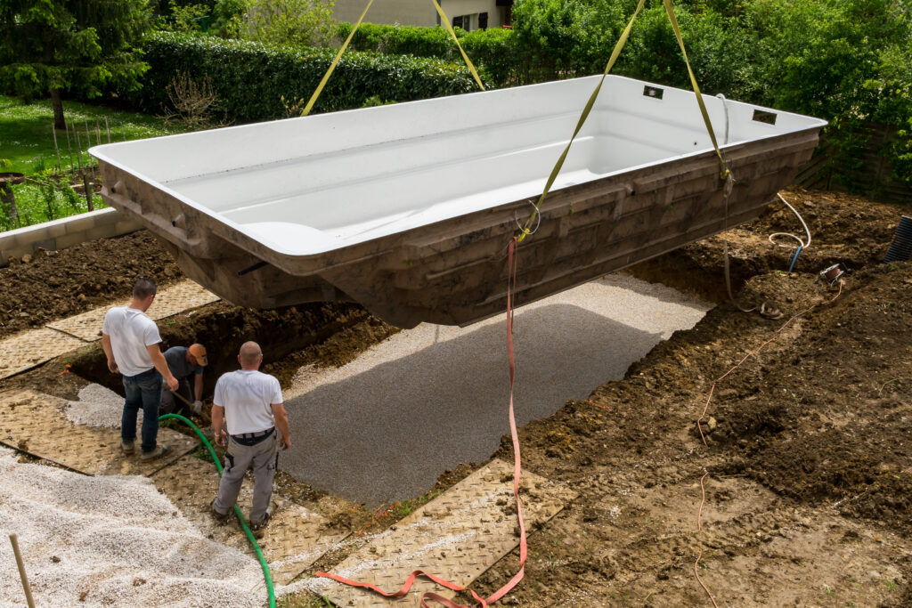 Fiberglass Inground Pool Installation Fiberglass inground pool shell being lowered by crane into an excavated backyard with gravel base and pool installation crew on site.