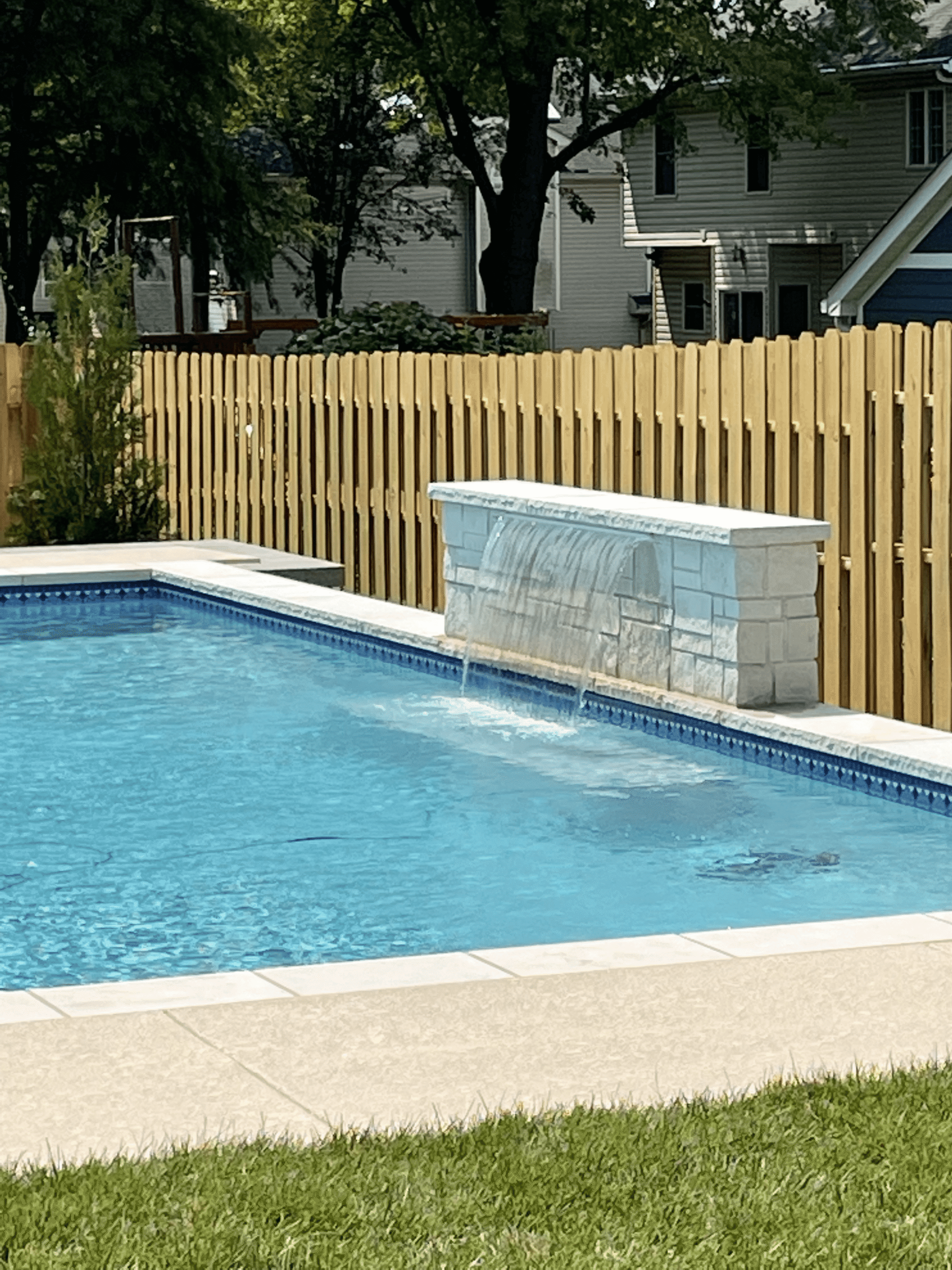 Inground Pool With Stone Waterfall Feature Inground swimming pool with a white stone waterfall feature, blue tile border, clear water, concrete pool deck, grass lawn, and privacy fence.