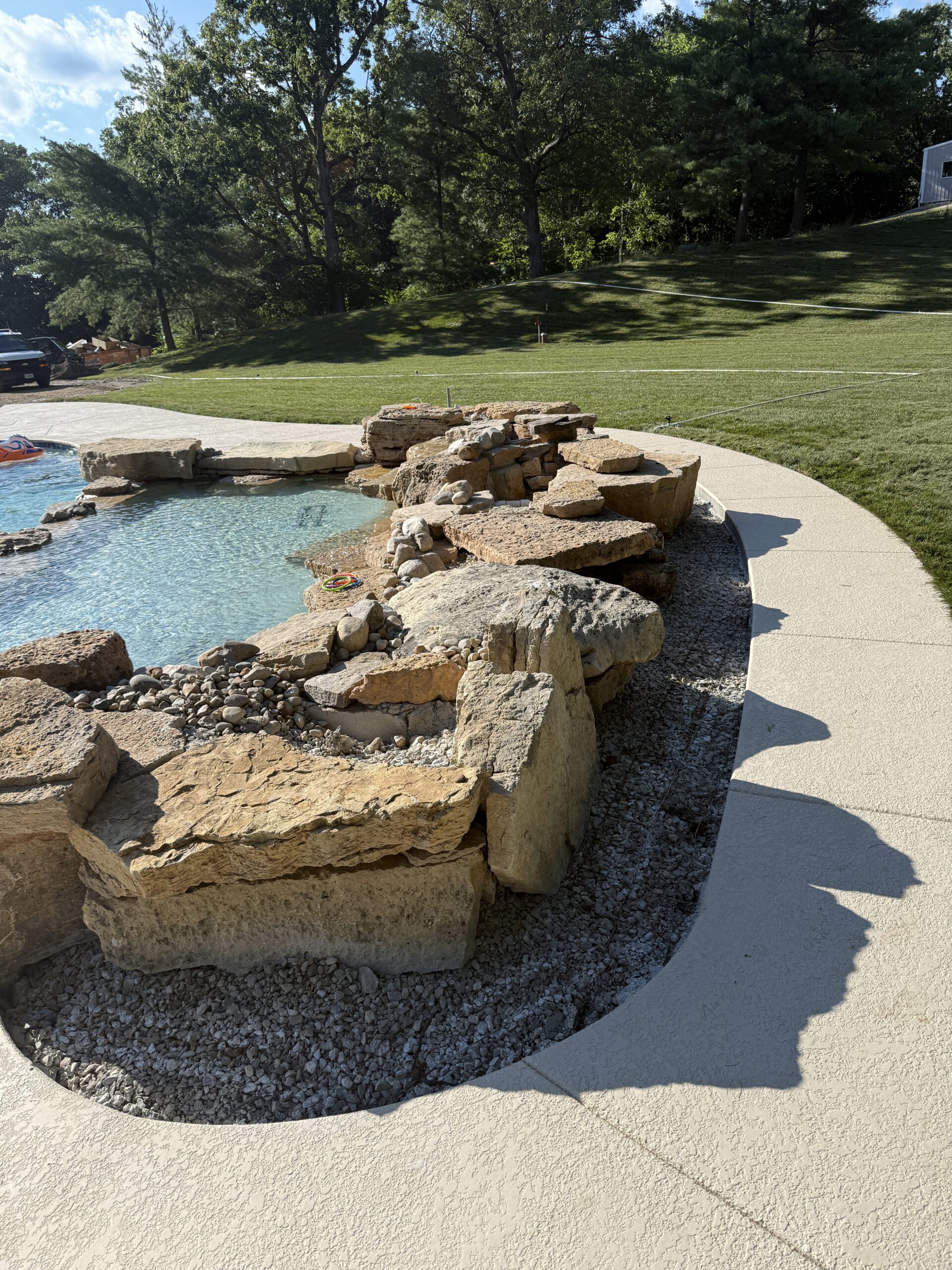 Custom Pool With Natural Stone Waterfall and Rock Landscaping Custom inground pool with natural stone waterfall, rock landscaping, curved concrete pool deck, clear blue water, and large backyard lawn.