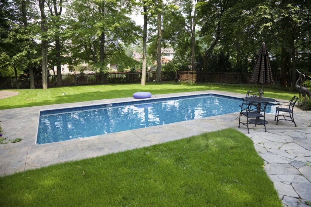 Backyard Inground Pool With Stone Patio Rectangular inground swimming pool with blue water, stone pool deck, green lawn, patio seating, umbrella, privacy fence, and mature trees in a backyard.